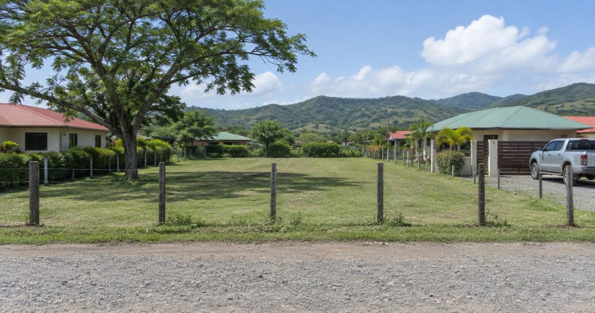 Lote plano con cerca de postes y alambre entre casas de lámina de color en Santa Cruz, Guanacaste, con cerros verdes al fondo
