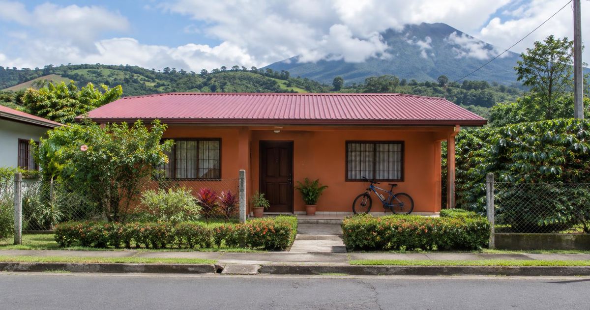 Casa de fachada terracota vista desde la acera, con jardín cuidado y cerros verdes al fondo, en la provincia de Alajuela